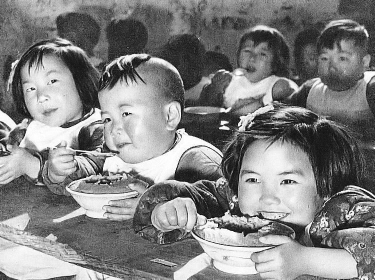 一群小孩子围着桌子捧着碗吃饭，笑得很开